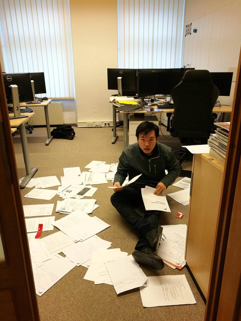 Mike sitzt von Dokumenten umgeben am Boden im Büro Hauptplatz und versucht Ordnung zu schaffen.