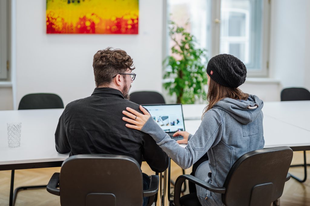 Zwei Personen sitzen zueinandergerichtet vor einem Laptop. Eine Person legt ihr Hand auf die Schulter der anderen.